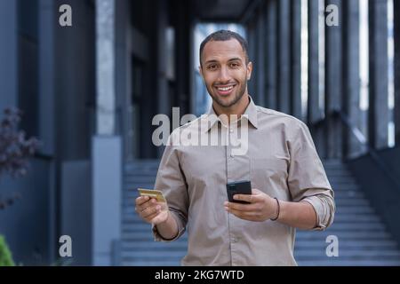 Ein junger afroamerikanischer Mann steht draußen. Er hat ein Telefon und eine Kreditkarte in der Hand, tätigt Online-Käufe, prüft das Konto, bezahlt die Bestellung. Er sieht in die Kamera und lächelt. Stockfoto