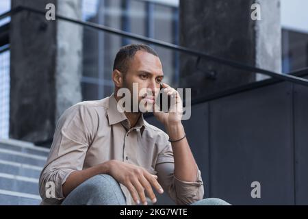 Porträt eines ernsten und schockierten hispanischen afroamerikaners. Er sitzt düster draußen auf der Treppe, spricht leider am Telefon, sieht weg. Stockfoto