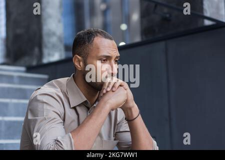 Porträt eines ernsten und traurigen hispanischen afroamerikanischen Mannes. Er sitzt draußen auf der Treppe, hält den Kopf in den Händen und schaut zur Seite. Stockfoto