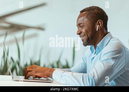 Lächelnder afrikanischer Geschäftsmann, der im modernen Büro auf einem Laptop sitzt Stockfoto
