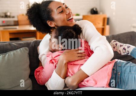 afroamerikanische glückliche Erwachsene Frau lächelt und umarmt ihr Töchtermädchen zu Hause Stockfoto