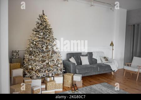 Stilvolles, trendiges skandinavisches Interieur mit einem riesigen schneebedeckten, luxuriösen, künstlichen Weihnachtsbaum in der Nähe eines grauen Sofas. Holzlaminat, Geschenke verpackt Stockfoto