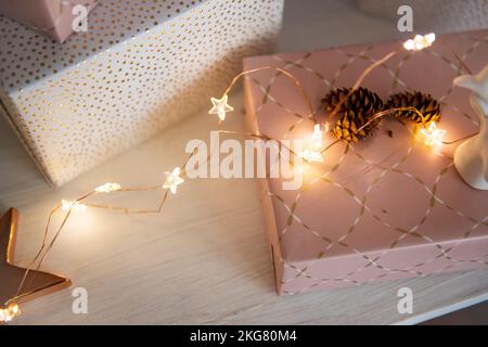 Weiße Rosenkästen mit Geschenken liegen auf einem Holztisch, mit einem leuchtend goldenen Stern, Girlande und natürlichen Kegeln. Festliche Wanddekoration. Postkarte zum Geburtstag Stockfoto