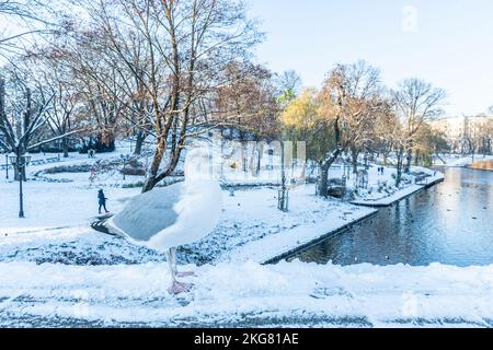 Winterlandschaft. Stadtpark mit Möwen im Vordergrund, Fluss, Brücke, Bänke und Bäume bedeckt vom Schnee, Menschen laufen in Riga, Lettland Stockfoto