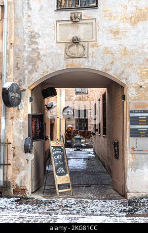 Eingang mit Schild für ein gemütliches Café in der Altstadt von Riga, Lettland, im Winter mit Tischen und Stühlen bedeckt von Schnee und altem Spotlight, vertikal Stockfoto