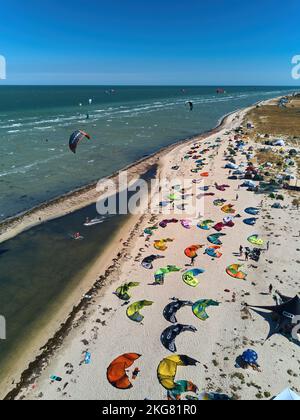 Blick von oben auf helle farbenfrohe Drachen, die am windigen Tag am Kitesurfplatz am Strand geparkt sind. Viele Fallschirme zum Kiteboarden liegen am Ufer Stockfoto