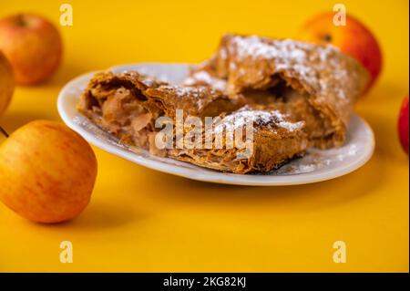 2 Stück Apfelkuchen (Strudel) auf weißem Teller, 4 roter Apfel auf gelbem Hintergrund, geschlossen. Stockfoto