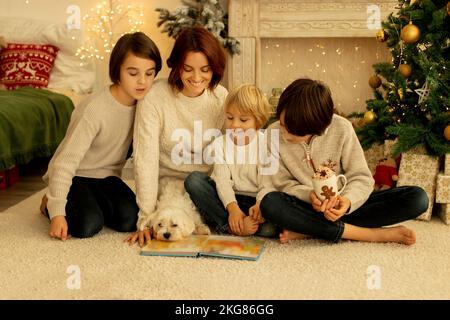 Mutter mit drei Kindern und Hund, zu Weihnachten zu Hause sitzen, gemütliche Atmosphäre, Weihnachtszeit ein Buch lesen Stockfoto