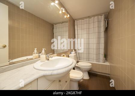 Kleines Badezimmer mit Porzellanwaschbecken mit weißer Kommode, langem Spiegel ohne Rahmen und Badewanne mit weißen Vorhängen Stockfoto