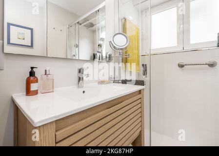 Kleines Badezimmer mit Porzellanwaschbecken auf Eichenschrank, Schiebedusche, rahmenlosem Spiegel und rundem Kosmetikspiegel Stockfoto