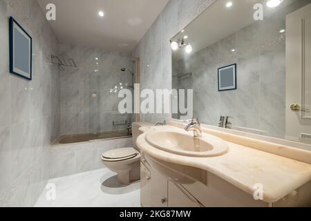 Klassisches Bad mit cremefarbenen Schränken mit farblich abgestimmter Ablagefläche, großem Spiegel an der Wand und cremefarbener Badewanne mit Leinwand Stockfoto