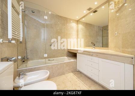 Badezimmer mit großen Spiegeln an den Wänden mit cremefarbenem Marmor und Böden aus demselben Material, weißen Holzmöbeln und Glasabtrennungen Stockfoto