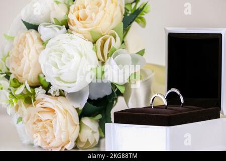 Ein paar exquisite glänzende Eheringe für ihn und sie, funkelnd in einer Ringbox, neben einem hübschen Strauß von Hochzeitsblumen, weißen und pfirsichfarbenen Rosen. Symbol Stockfoto