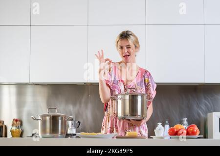 Eine Frau mit Kochtopf steht neben dem Tisch in der Küche zu Hause. Die Hausfrau kocht morgens in der Küche. Stockfoto