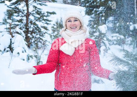 Eine Frau mittleren Alters lacht, wenn sie unter dem Schneefall von Bäumen steht Stockfoto