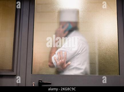 Silhouette hinter dem Glas eines Mannes in einer Koch-Uniform, der am Telefon spricht Stockfoto