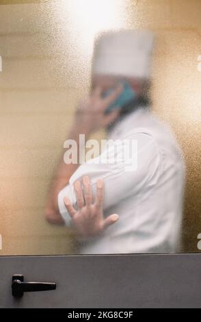 Silhouette hinter dem Glas eines Mannes in einer Koch-Uniform, der am Telefon spricht Stockfoto