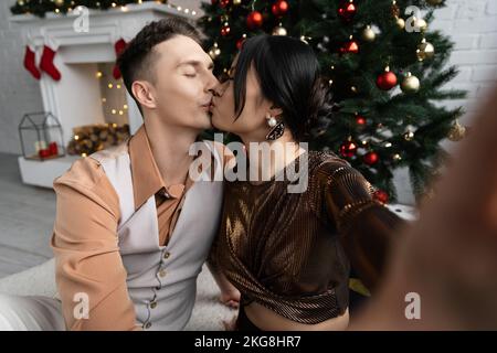 Interracial Paar mit geschlossenen Augen küssen, während unter Weihnachtsbaum sitzen Stockfoto