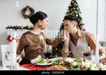 Tätowierte Mann küsst Hand der asiatischen Frau in elegantem Outfit, während mit romantischen Abendessen an Weihnachten zu Hause Stockfoto