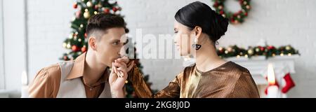 Junger Mann küsst die Hand der eleganten asiatischen Frau im Wohnzimmer während der Weihnachtsfeier, Banner Stockfoto