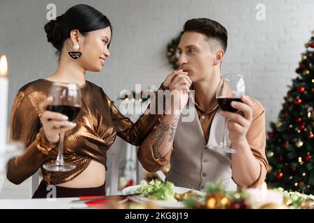 Tätowierte Mann küsst Hand von eleganten asiatischen Frau, während Weihnachten zu Hause feiern Stockfoto