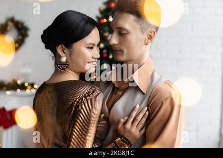 Brunette asiatische Frau in festlichen Kleidern lächelt in der Nähe jungen Mann während der Weihnachtsfeier zu Hause Stockfoto