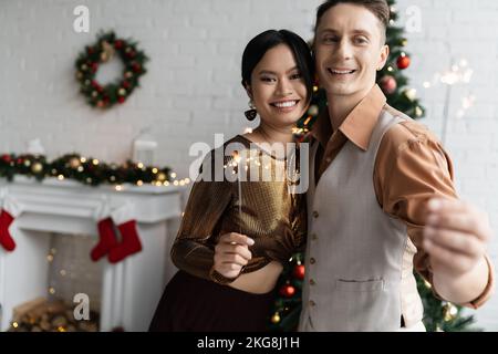 Fröhliches und elegantes interrassisches Paar mit glänzenden Wunderkerzen und Blick auf die Kamera während der Weihnachtsfeier Stockfoto