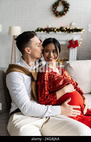 Mann küsst schwanger asiatische Frau und umarmt ihren Bauch während der Weihnachtsfeier Stockfoto