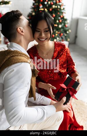 Junger Mann hält Weihnachtsgeschenk und küsst schwanger asiatische Frau in eleganten Kleidern Stockfoto