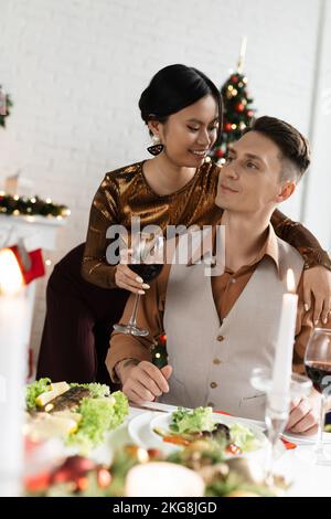 Glückliche asiatische Frau mit einem Glas Wein und umarmte den Mann in der Nähe des festlichen Abendessens während der Weihnachtsfeier Stockfoto