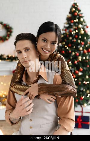 Fröhliche asiatische Frau umarmt Ehemann in der Nähe geschmückten Weihnachtsbaum Stockfoto