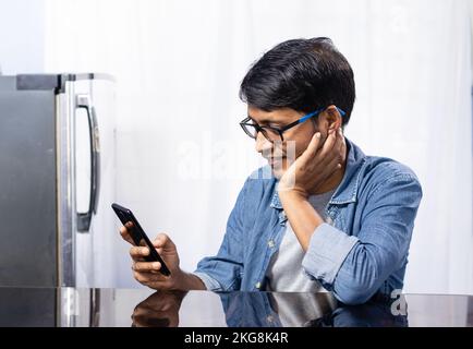 Ein indischer Mann, der mit einem lächelnden Gesicht zu Hause auf sein Smartphone schaut Stockfoto