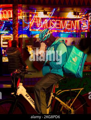 Ein Mann in einer Auslieferungsuniform, der eine Umhängetasche in einem Fahrradhelm trug, Fahrrad fuhr und sein Handy in London anschaute. Stockfoto