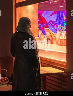 Eine alleinstehende Frau mit weißem Kopftuch und braunem Mantel blickt durch ein Fenster in einen Laden, der von bunten Neonlichtern beleuchtet wird. Stockfoto