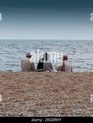 Eine Familie saß an einem steinernen Kieselstrand unter einem grauen Himmel Stockfoto