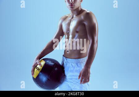 Zugeschnittenes Bild eines jungen Muskelmannes, der sich mit einem über blauem Hintergrund isolierten Medizinball in Neonlicht posiert Stockfoto