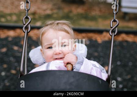 Eine Nahaufnahme eines niedlichen 1-jährigen Mädchens auf einer Schaukel mit einem Kiefernzapfen. Morris Plains, NJ, November. Stockfoto