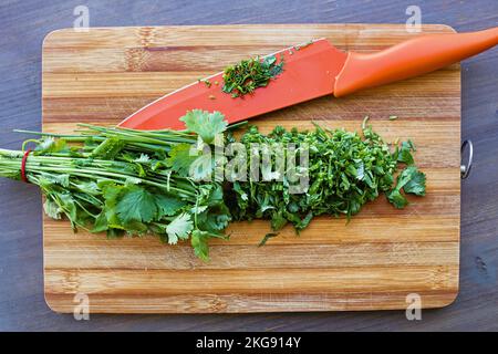 Ein Holzbrett mit einem großen Haufen Kräuter, frisch in zwei Hälften geschnitten. Daneben ist ein orangefarbenes, großes Messer mit Kräuterresten auf der breiten Klinge. Obere V-Achse Stockfoto