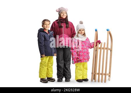 Zwei Mädchen und ein Junge in Winterkleidung stehen mit einem Holzschlitten isoliert auf weißem Hintergrund Stockfoto