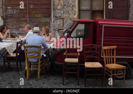 Rom, Italien: Paare speisen an kleinen Tischen in einer kopfsteingepflasterten Straße in Trastevere im Freien. Holzstühle vor einem kleinen roten Truck, einem Piaggio Ape 50. Stockfoto