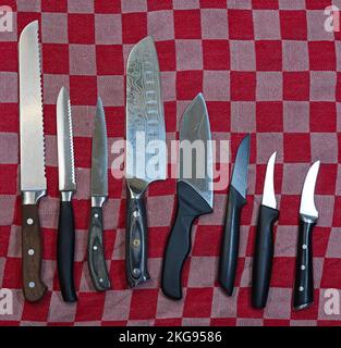 Acht Küchenmesser auf einem rot-weißen karierten Handtuch. Das erste ist ein Brotmesser. Als nächstes ein Tomatenmesser. Das vierte ist ein japanisches Gyuto-Messer. Stockfoto