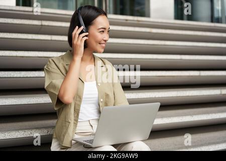 Porträt einer stilvollen jungen Frau, die lächelt, mit Kopfhörern und Laptop auf der Treppe sitzt, Hausaufgaben für die Universität macht Stockfoto