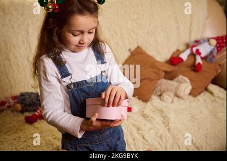 Charmantes kleines Mädchen mit Hirschgeweih-Reifen, packt wunderschöne herzförmige rosa Geschenkbox aus. Der Zweite Weihnachtsfeiertag. Weihnachtszeit Stockfoto