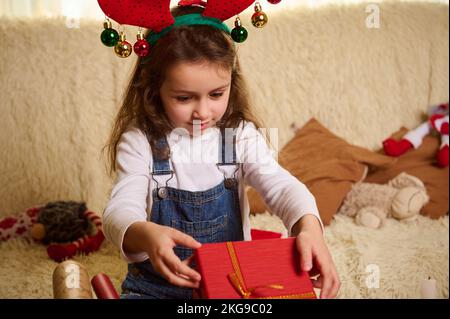 Ein bezauberndes, kaukasisches Mädchen, das ein Hirschgeweih trägt und eine wunderschöne rote Geschenkbox auspackt. Der Zweite Weihnachtsfeiertag. Weihnachtszeit Stockfoto