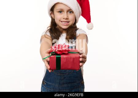 Ein hübsches Mädchen mit Weihnachtsmann-Hut, ein glückliches Geschenk mit roter Schleife, ein süßes Lächeln in die Kamera. Heiligabend Stockfoto