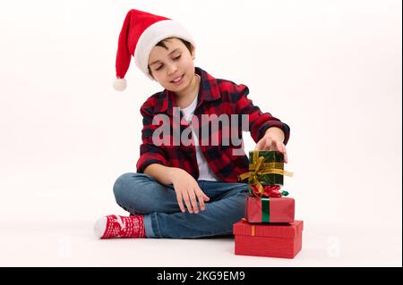 Ein hübscher Junge mit Weihnachtsmannmütze und kariertem Hemd, stapelt Weihnachtsgeschenke, sitzt auf weißem Hintergrund. Neujahrsfeier Stockfoto