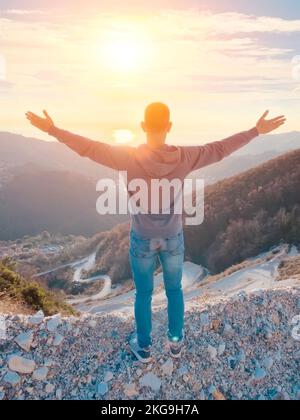 Ein Mann steht mit erhobenen Händen auf einem Berg. Im Hintergrund, Sonnenuntergang und Meer, gewundene Serpentine Road unten. Das Konzept des freien Reisens. Stockfoto