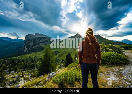 Beschreibung: Sportliche Frau in Outdoor-Jacke sieht sich die wunderschöne Bergkulisse bei Sonnenuntergang an. Falzarego Pass, Dolomiten, Südtirol, Italien, Europa. Stockfoto