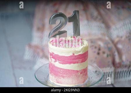 21. Geburtstagskuchen, rosa und silberne Kuchen mit ein paar Streuseln und 21. Kerzenlicht mit einem Frauenkleid im Hintergrund. Stockfoto