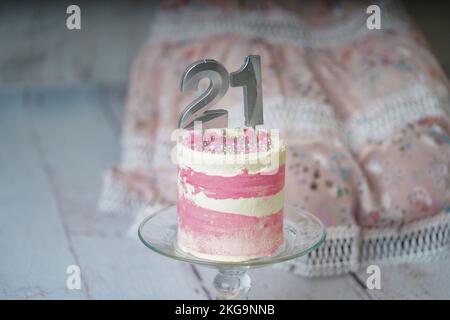 21. Geburtstagskuchen, rosa und silberne Kuchen mit ein paar Streuseln und 21. Kerzenlicht mit einem Frauenkleid im Hintergrund. Stockfoto